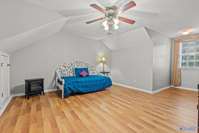 bedroom featuring vaulted ceiling, a textured ceiling, a wood stove, ceiling fan, and light hardwood / wood-style floors