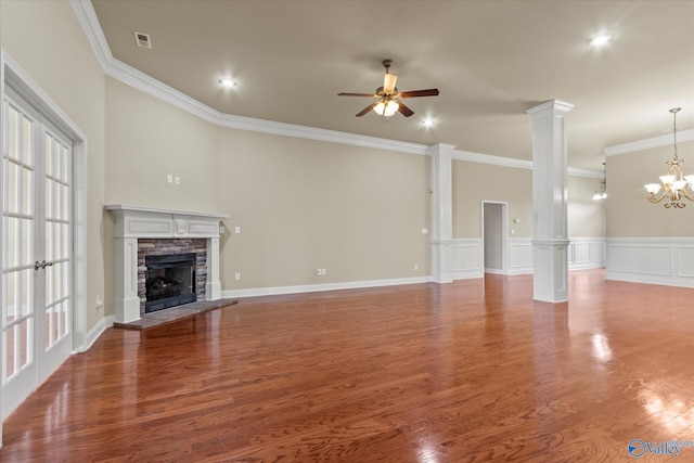 unfurnished living room featuring decorative columns, ceiling fan with notable chandelier, crown molding, wood-type flooring, and a stone fireplace