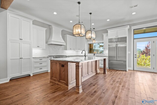 kitchen featuring premium range hood, a center island with sink, white cabinets, and stainless steel built in refrigerator