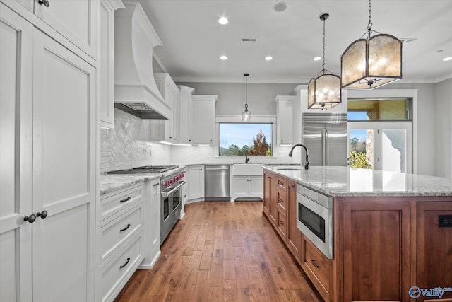 kitchen with hanging light fixtures, light stone counters, built in appliances, a kitchen island with sink, and white cabinets