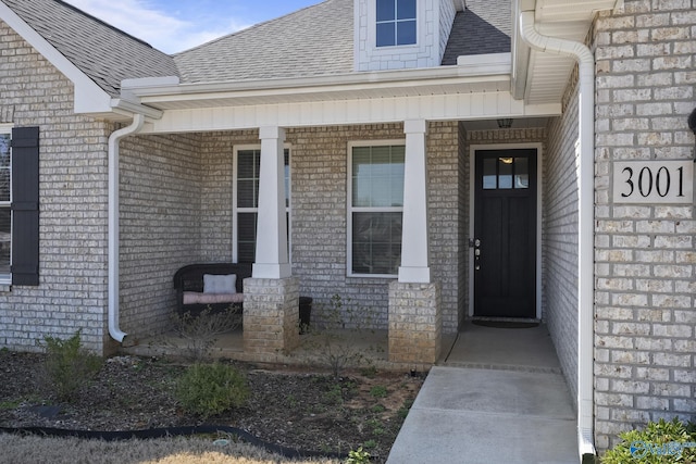 doorway to property featuring covered porch, roof with shingles, and brick siding