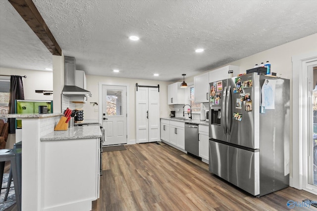 kitchen with a barn door, stainless steel appliances, white cabinetry, wall chimney range hood, and dark wood finished floors