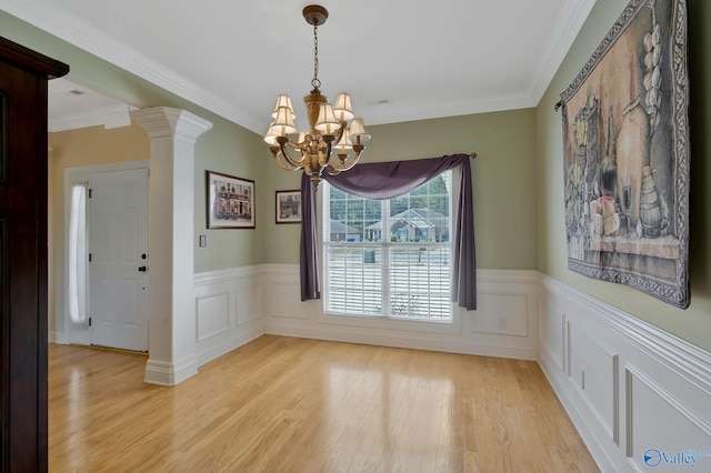 unfurnished dining area with crown molding, a chandelier, a wainscoted wall, light wood-type flooring, and decorative columns