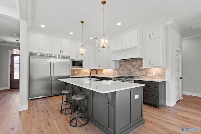 kitchen featuring built in appliances, decorative light fixtures, an island with sink, and white cabinets
