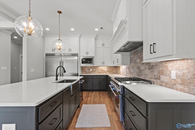 kitchen with custom exhaust hood, white cabinetry, built in appliances, hanging light fixtures, and a center island with sink