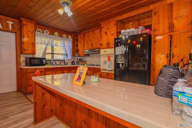 kitchen with tile countertops, wooden ceiling, backsplash, black appliances, and light hardwood / wood-style flooring