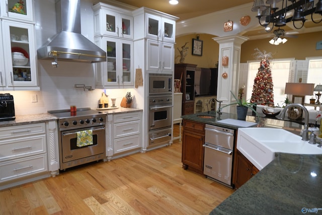 kitchen with sink, wall chimney range hood, dark stone counters, white cabinets, and appliances with stainless steel finishes