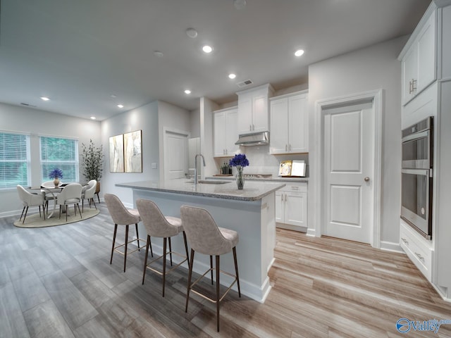 kitchen featuring a kitchen island with sink, under cabinet range hood, a sink, a kitchen breakfast bar, and white cabinets