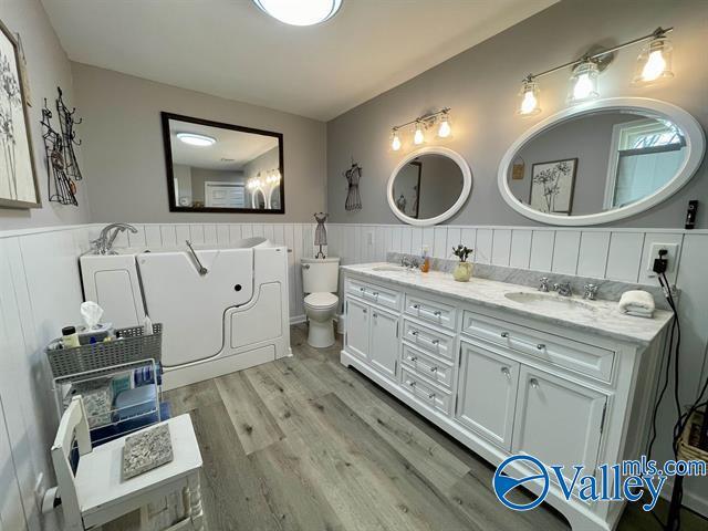 full bath with wood finished floors, double vanity, a sink, and a wainscoted wall