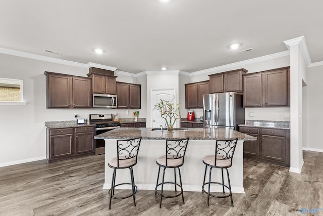 kitchen featuring dark hardwood / wood-style flooring, dark brown cabinets, appliances with stainless steel finishes, a center island with sink, and ornamental molding