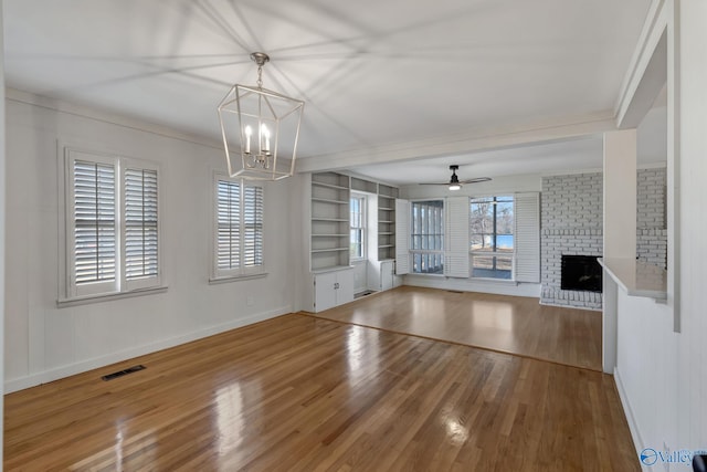 unfurnished living room with built in shelves, visible vents, a brick fireplace, wood finished floors, and ceiling fan with notable chandelier