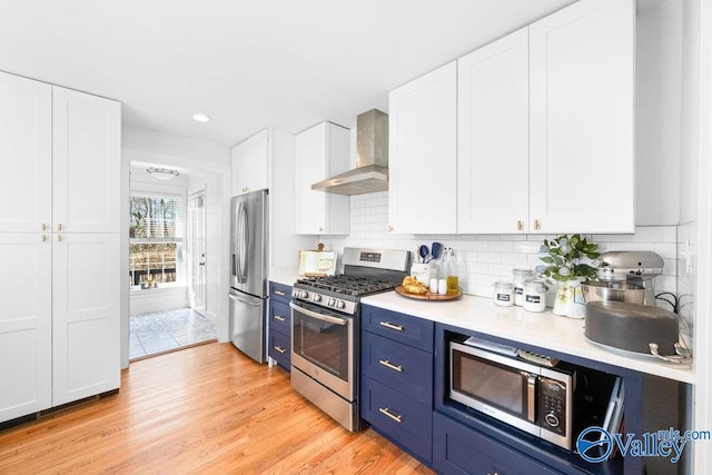 kitchen featuring white cabinetry, stainless steel appliances, blue cabinets, and wall chimney range hood