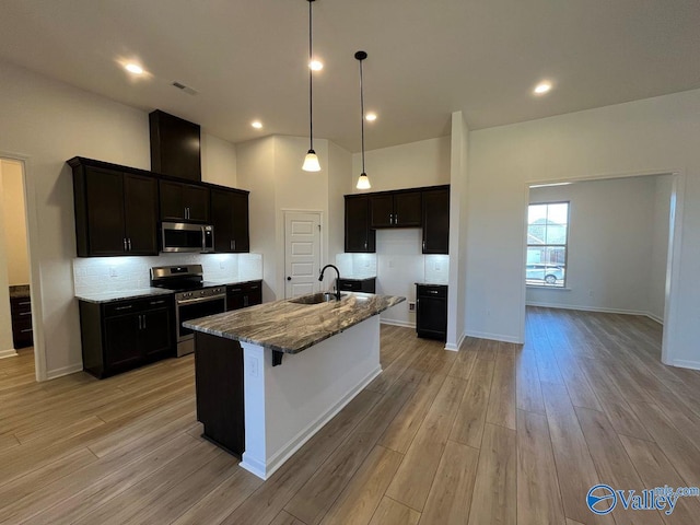 kitchen featuring stainless steel appliances, sink, light wood-type flooring, light stone counters, and a center island with sink