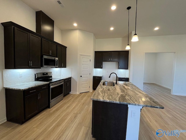 kitchen with an island with sink, stainless steel appliances, light stone countertops, light hardwood / wood-style flooring, and sink