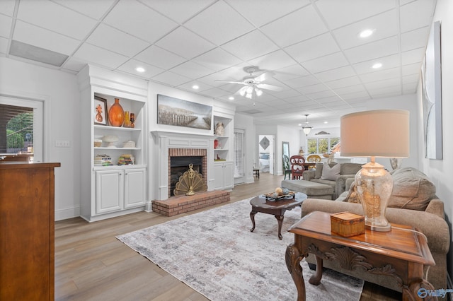 living room featuring light wood-style flooring, ceiling fan, a brick fireplace, baseboards, and a drop ceiling