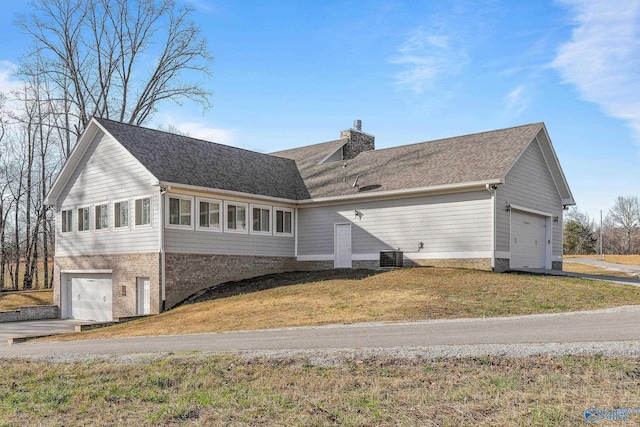 exterior space with concrete driveway, roof with shingles, central air condition unit, a chimney, and a yard