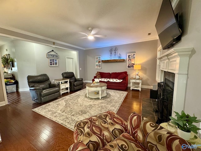 living area with dark wood-style flooring, a tiled fireplace, baseboards, a ceiling fan, and ornamental molding