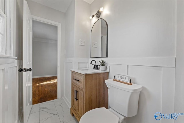 bathroom featuring toilet, a wainscoted wall, marble finish floor, vanity, and a decorative wall