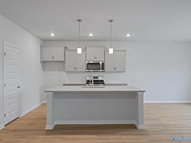 kitchen featuring light stone counters, pendant lighting, a kitchen island with sink, appliances with stainless steel finishes, and light wood-type flooring