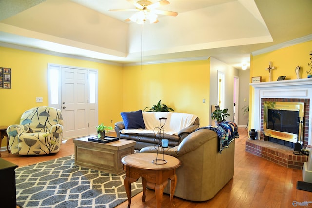 living room featuring a brick fireplace, hardwood / wood-style floors, a tray ceiling, and ceiling fan