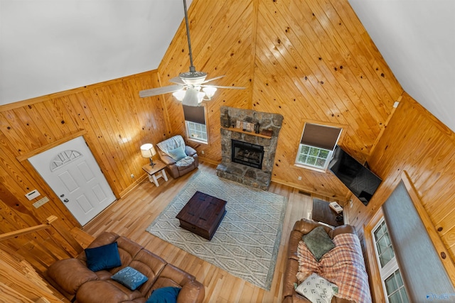 living room featuring wood walls, a stone fireplace, ceiling fan, hardwood / wood-style flooring, and a high ceiling