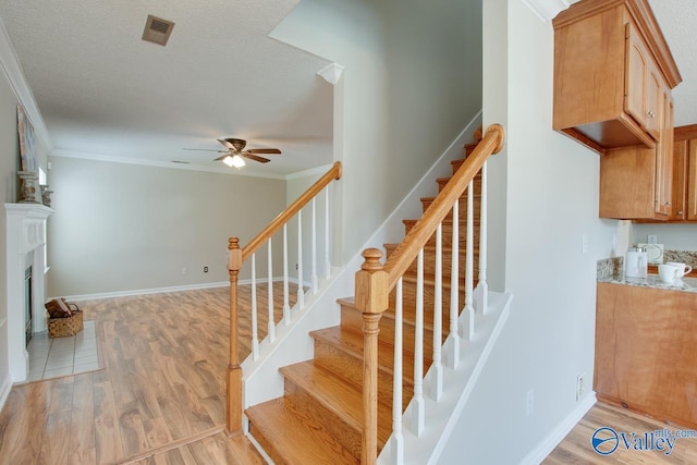 staircase with a ceiling fan, wood finished floors, ornamental molding, a textured ceiling, and a fireplace