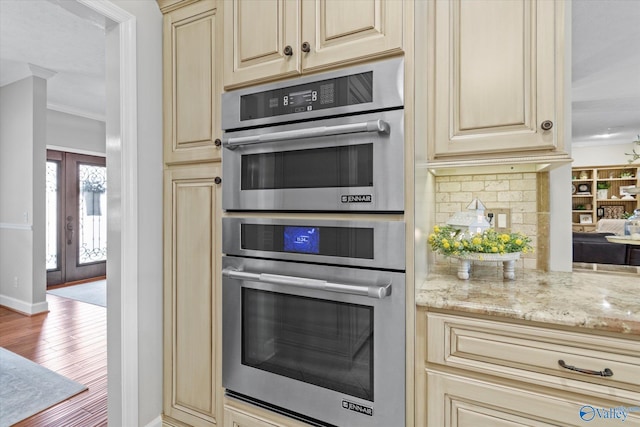 kitchen featuring tasteful backsplash, wood-type flooring, stainless steel double oven, light stone countertops, and cream cabinetry