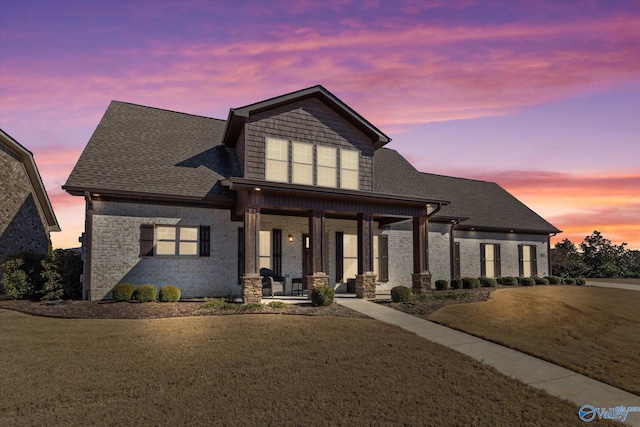 view of front of house featuring a porch, roof with shingles, a yard, and brick siding