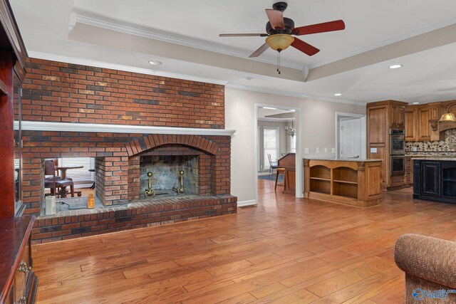 living area featuring crown molding, a tray ceiling, a fireplace, light wood-style floors, and a ceiling fan