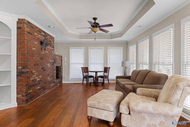 living area featuring built in shelves, a ceiling fan, a fireplace, wood-type flooring, and a raised ceiling