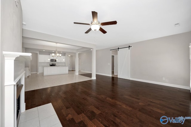 unfurnished living room with a barn door, visible vents, baseboards, light wood-style flooring, and ceiling fan with notable chandelier