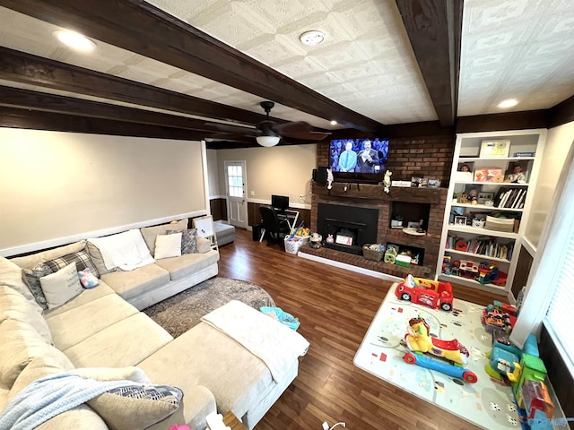 living area with a wainscoted wall, wood finished floors, a ceiling fan, a brick fireplace, and beamed ceiling