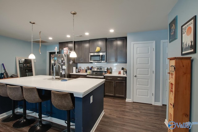 kitchen with a center island with sink, stainless steel appliances, decorative light fixtures, sink, and dark wood-type flooring