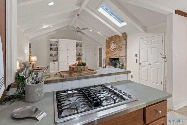 kitchen with vaulted ceiling with skylight, stainless steel gas stovetop, a ceiling fan, a stone fireplace, and white cabinets