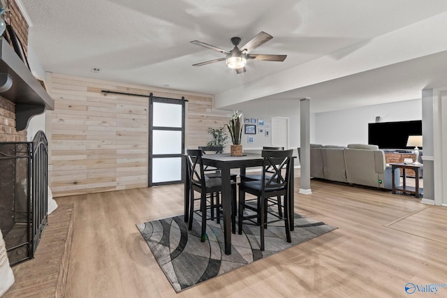 dining area featuring a barn door, wood walls, ceiling fan, light wood finished floors, and a brick fireplace
