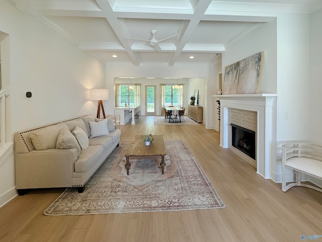 living room featuring light wood-type flooring, beam ceiling, coffered ceiling, a brick fireplace, and ceiling fan