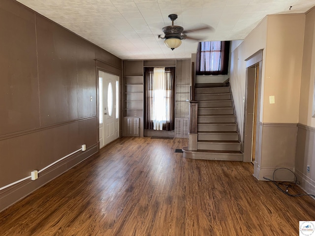 unfurnished living room with stairs, ceiling fan, a wainscoted wall, and dark wood finished floors