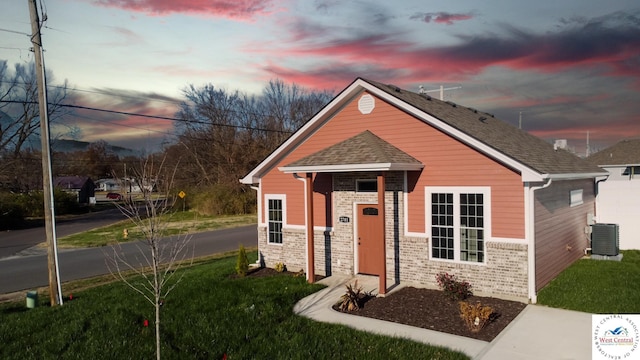 view of front of property featuring central AC, brick siding, roof with shingles, and a lawn