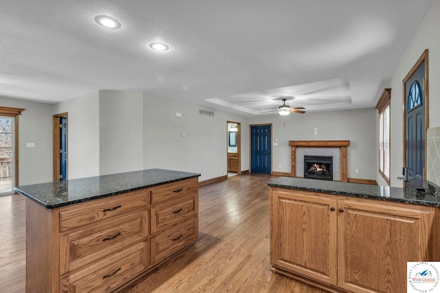 kitchen with light wood finished floors, a raised ceiling, visible vents, a tiled fireplace, and dark stone countertops