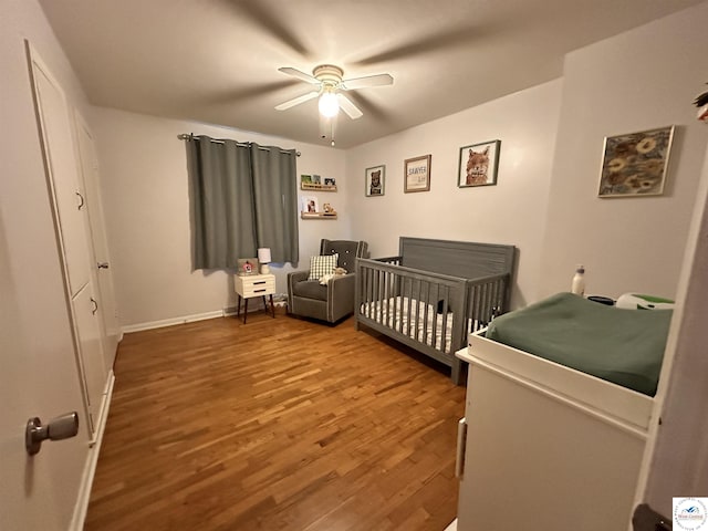 bedroom featuring a nursery area, ceiling fan, wood finished floors, and baseboards