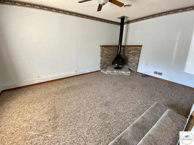 unfurnished living room with visible vents, a wood stove, carpet flooring, ceiling fan, and baseboards