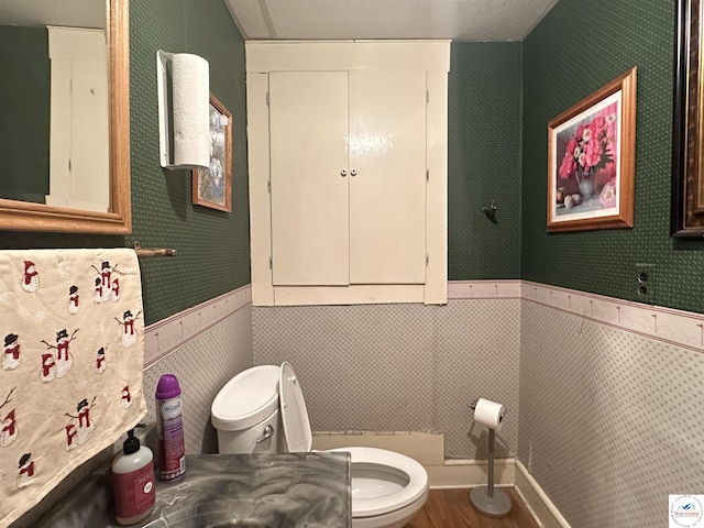 half bath with wallpapered walls, toilet, wood finished floors, and wainscoting