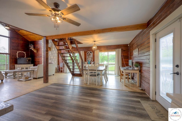 unfurnished dining area featuring lofted ceiling with beams, a ceiling fan, wooden walls, wood finished floors, and stairs