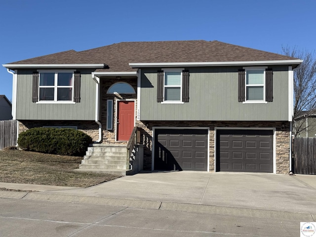 split foyer home with driveway, stone siding, roof with shingles, an attached garage, and fence