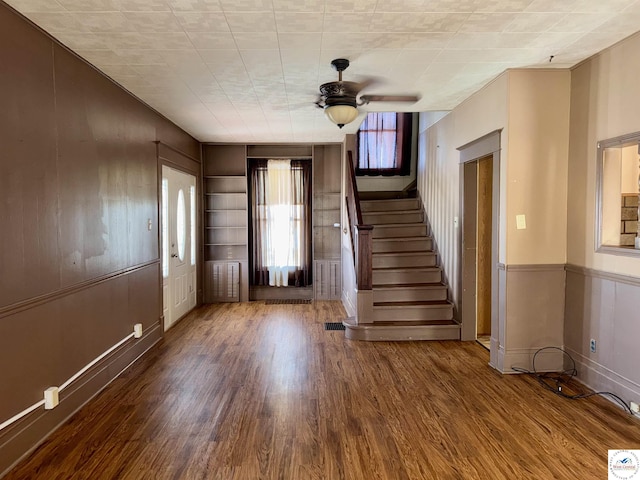 unfurnished living room with a ceiling fan, stairway, wood walls, and wood finished floors