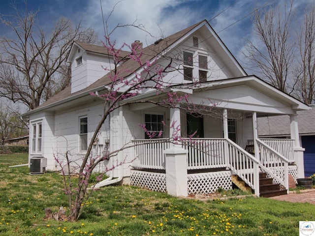 view of front of home with a chimney, covered porch, central air condition unit, roof with shingles, and a front yard