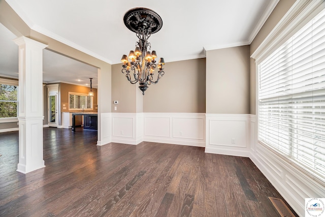 unfurnished dining area featuring decorative columns, a chandelier, dark wood-type flooring, a wainscoted wall, and crown molding