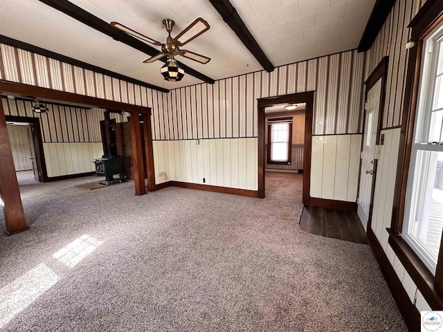 empty room with a wood stove, ceiling fan, carpet flooring, and beamed ceiling
