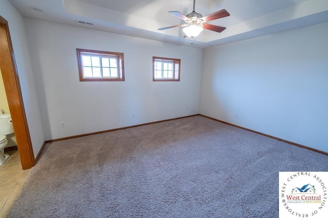 empty room with a ceiling fan, light carpet, a raised ceiling, and baseboards