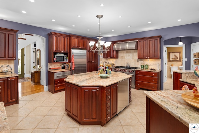 kitchen with decorative backsplash, light tile patterned floors, high end appliances, wall chimney exhaust hood, and arched walkways